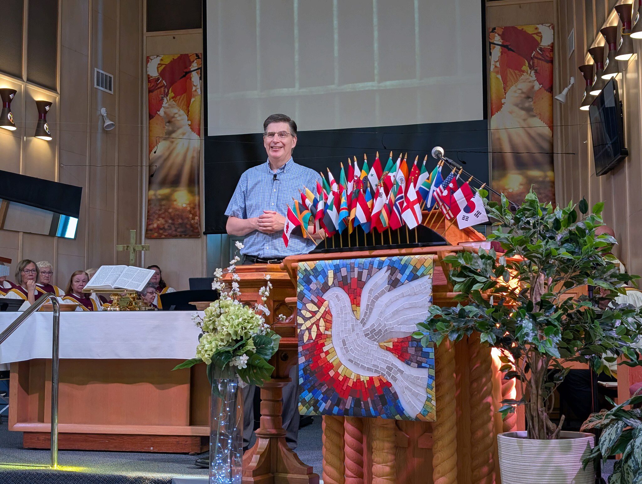 Pastor Greg standing in the sanctuary by the pulpit. His hands are folded across his blue checkered shirt.