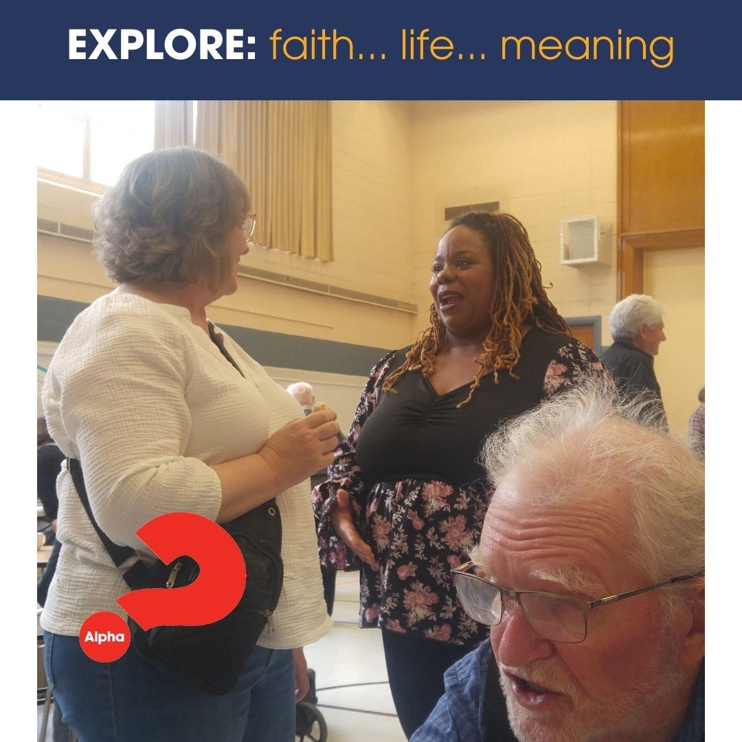 Two women are standing in a church gymnasium, having a friendly conversation. At the bottom of the picture, is the face of a male, who is talking to someone off camera. 